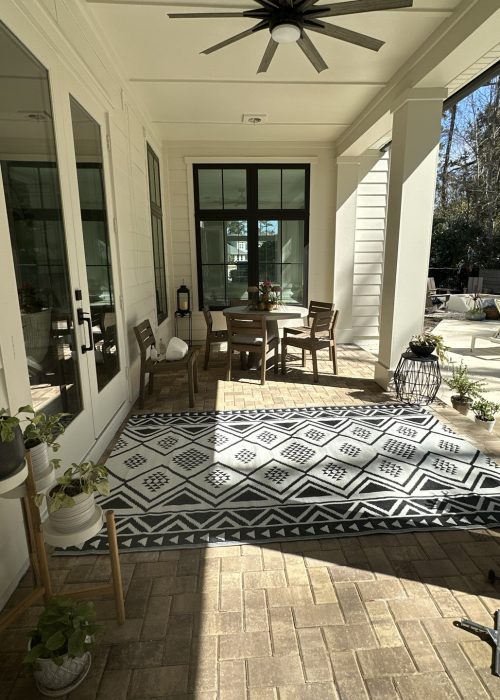 A welcoming covered porch featuring a round table with four chairs, a patterned outdoor rug, and potted plants, perfect for relaxing or entertaining guests in a charming residential setting.