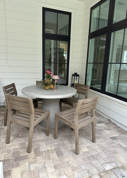 Cozy outdoor dining space on a screened porch with a round table, four wooden chairs, and decorative flowers, surrounded by large windows and white siding.