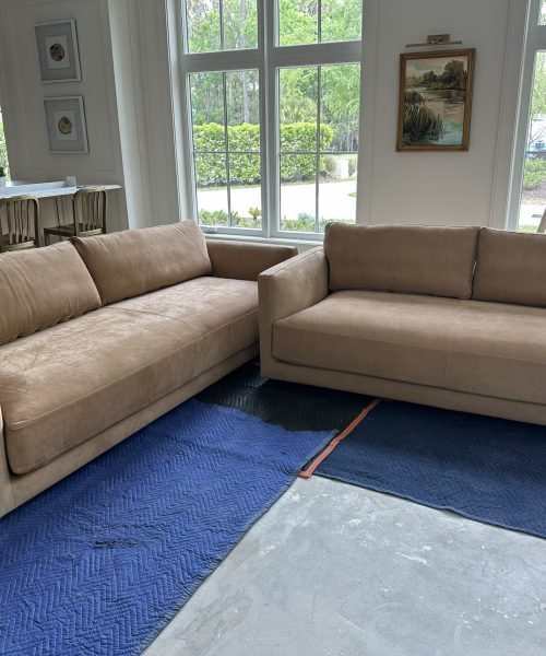 Spacious living room featuring two beige sofas, large windows with greenery outside, and artwork on the walls, creating a bright and inviting atmosphere.