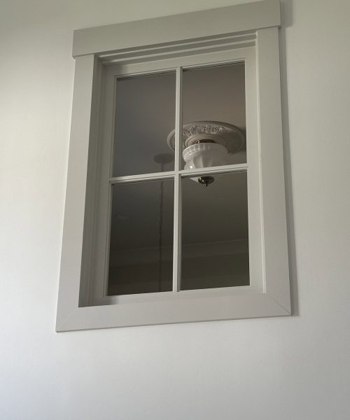 Bright white window with four panes and decorative trim, set against a light-colored wall. The window offers a view of a ceiling fan and light fixture inside the room.