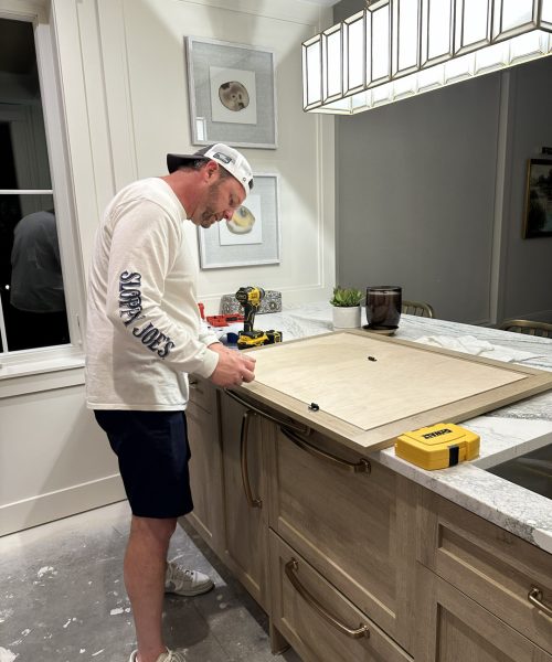 A man working on a kitchen countertop renovation, measuring and preparing the surface with tools, in a modern, well-lit kitchen with white cabinets and decorative wall art.