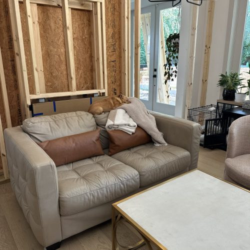Cozy living room with a beige leather tufted sofa, featuring brown and neutral pillows and a blanket, under construction with exposed wooden wall studs and a modern black chandelier, natural light coming in.