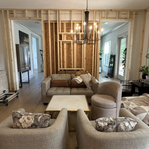 Modern living room with unfinished wall framing, featuring a beige sofa, floral armchairs, a round side chair, a square coffee table, and a black chandelier, highlighting a home renovation or construction project.