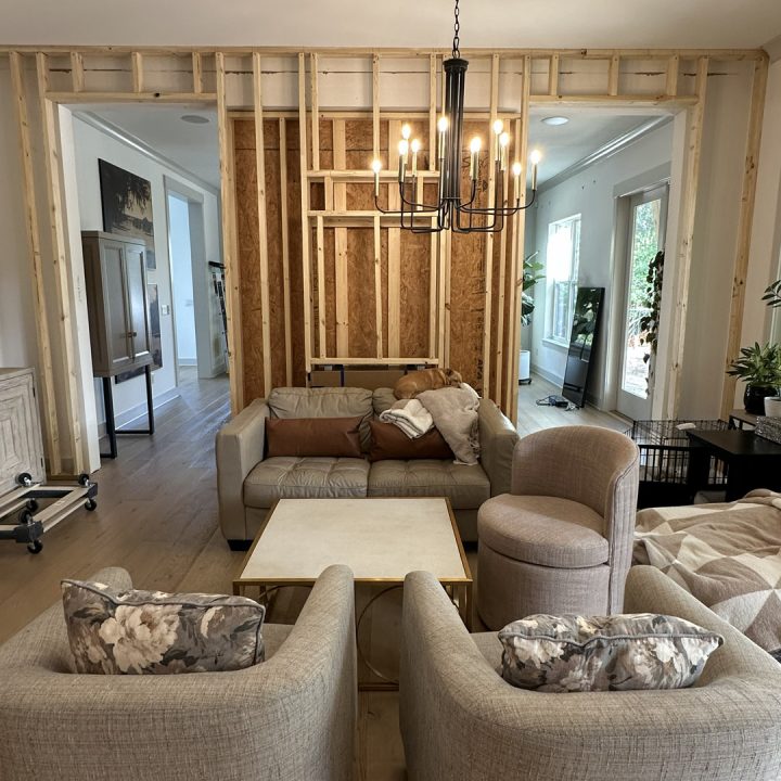 Modern living room with unfinished wall framing, featuring a beige sofa, floral armchairs, a round side chair, a square coffee table, and a black chandelier, highlighting a home renovation or construction project.