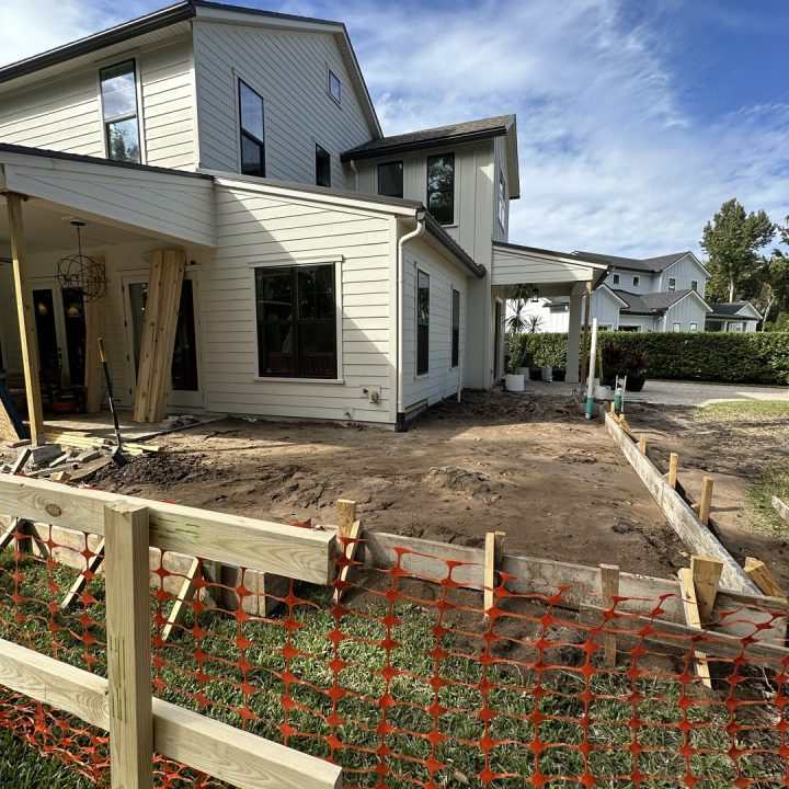 Newly constructed white modern house with multiple stories under construction, featuring large windows and a sloped roof, surrounded by a landscaped yard and construction materials.