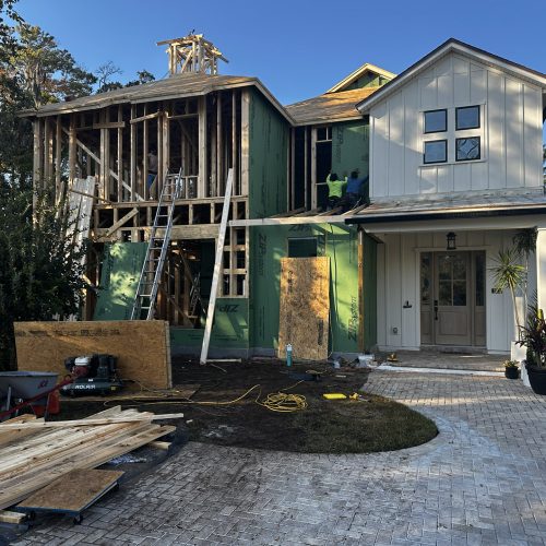 New house under construction with a partly built second story, wooden framework, and workers installing exterior siding; driveway, plants, and construction tools visible.