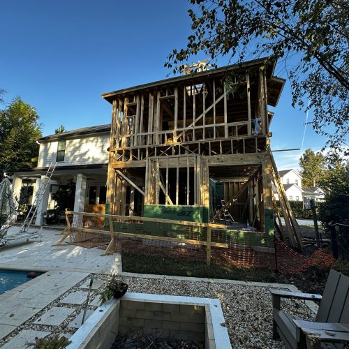 New home construction with a partially built multi-story wooden frame, situated next to a finished white house, under a bright blue sky, with surrounding trees and a backyard pool area.