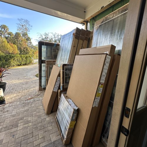 New home construction materials including windows, doors, and framing supplies stacked at the entrance of a house under construction in a sunny outdoor setting.