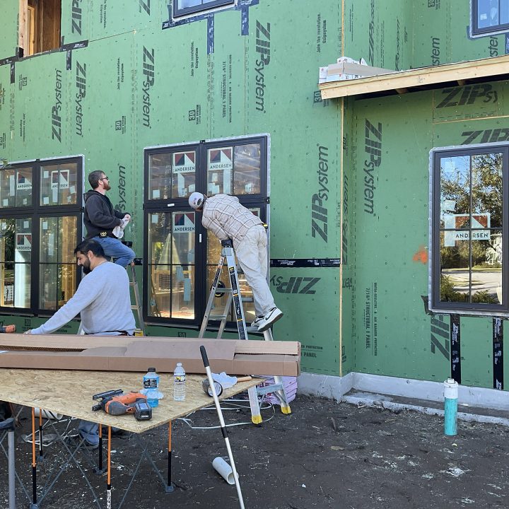 Newly constructed home with green exterior sheathing, featuring workers installing black-framed windows on a sunny day.