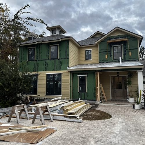 1. New two-story house under construction with green exterior wrapping, beige siding, and a front porch, set against a cloudy sky.