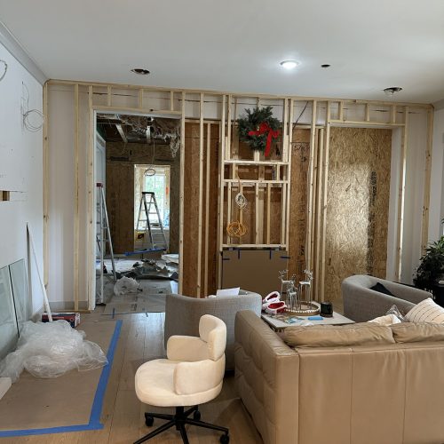Newly constructed living room with exposed wall framing during renovation, featuring holiday decor with a wreath and red bow, and modern furniture, illustrating ongoing home building or remodeling process.