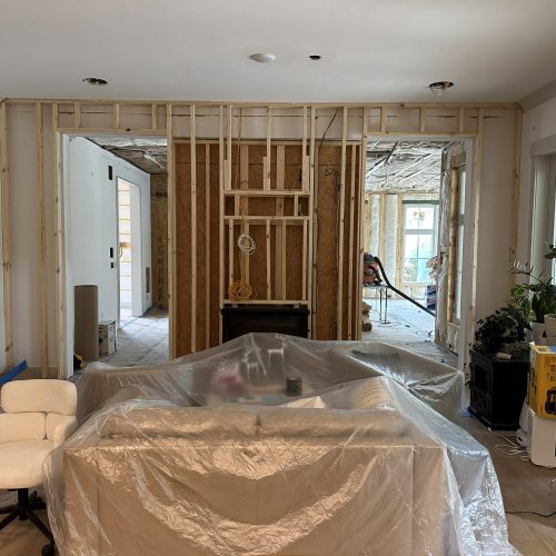 Newly renovated living room under construction with exposed wall framing, construction supplies, and a plastic-covered sofa, showcasing ongoing home remodeling by John Merrill Homes.