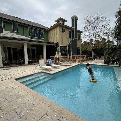 Bright blue swimming pool with steps, surrounded by a beige stone deck, adjacent to a modern home with outdoor seating, under an overcast sky.