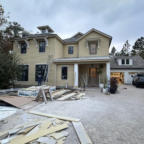 Newly constructed yellow farmhouse-style home under construction with workers, tools, and building materials in a suburban neighborhood with a paved driveway and surrounding trees.