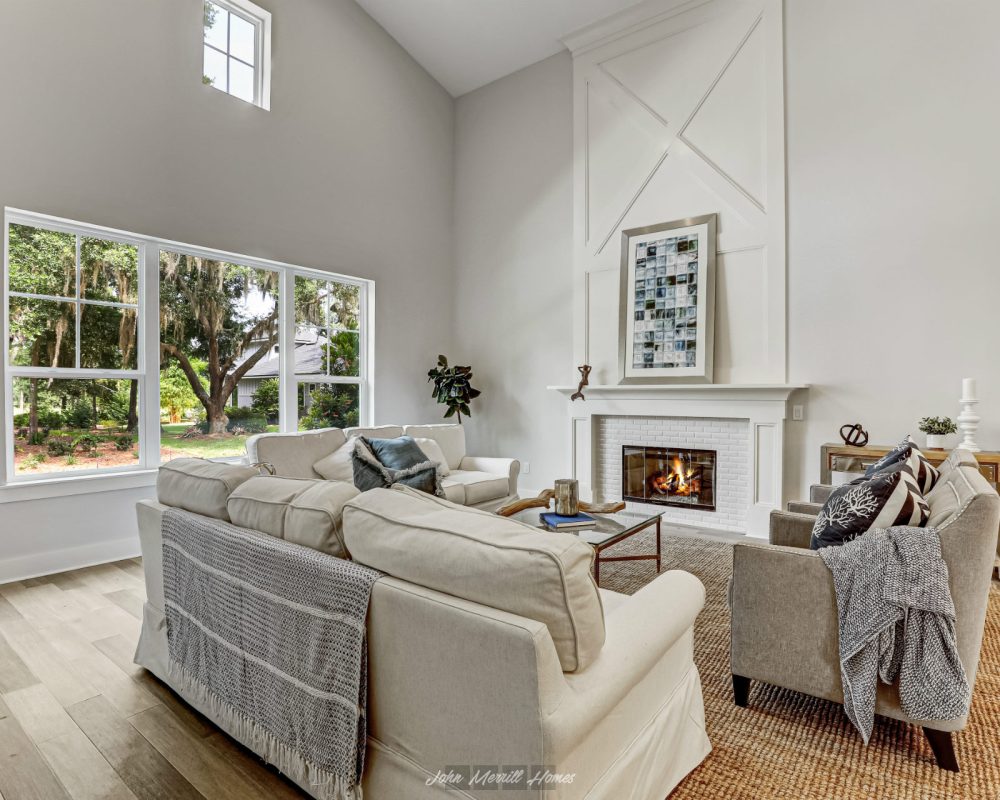 Modern living room with a cozy fireplace, large windows showcasing lush outdoor greenery, and stylish furniture creating an inviting atmosphere | Bright, airy open-concept space with high ceilings, white walls, and natural light | Image of a contemporary home interior emphasizing comfort and elegance, perfect for relaxing or entertaining guests.