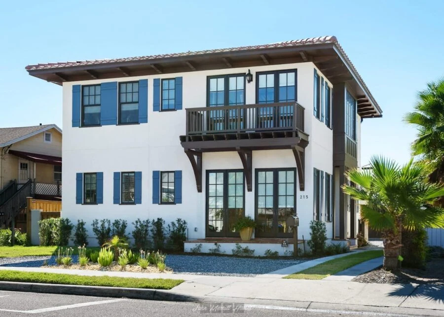 A modern coastal-style two-story house featuring a balcony, large windows, and well-maintained landscaping with palm trees and shrubs.