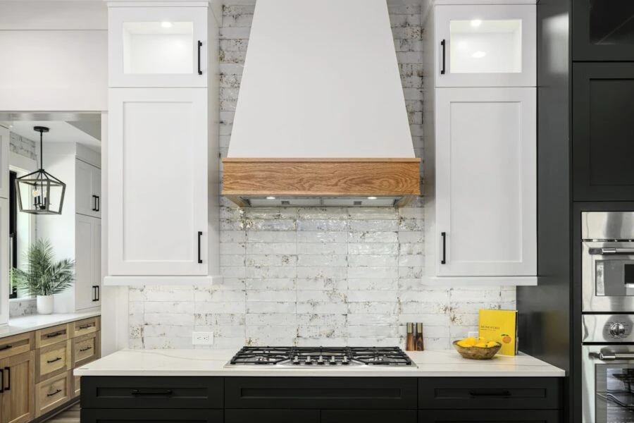 Contemporary kitchen featuring white cabinetry, black lower cabinets, a white brick backsplash, and a wooden range hood, creating a sleek and stylish cooking space.