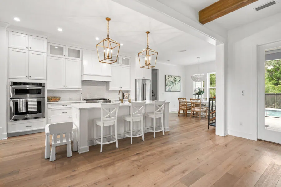 Bright modern white kitchen with stainless steel appliances, gold pendant lights, and a spacious open floor plan with dining area and large windows showcasing greenery.