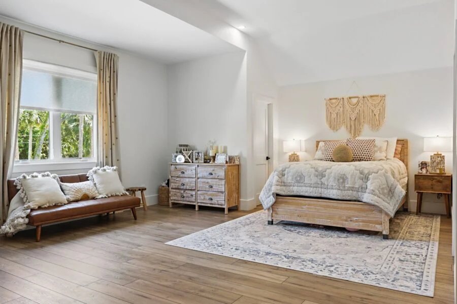 Bright and airy bedroom featuring a large bed with decorative pillows, a cozy sitting area with a leather bench, and natural light streaming through windows with beige curtains.