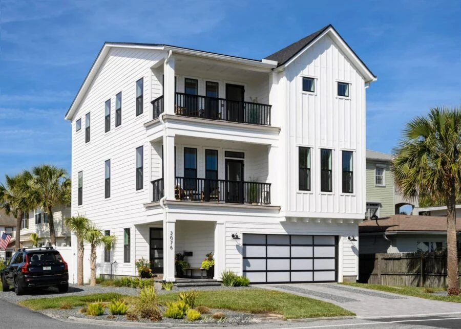 A contemporary white house featuring three levels, black balcony railings, and large windows, situated in a sunny neighborhood with palm trees and a well-maintained lawn.